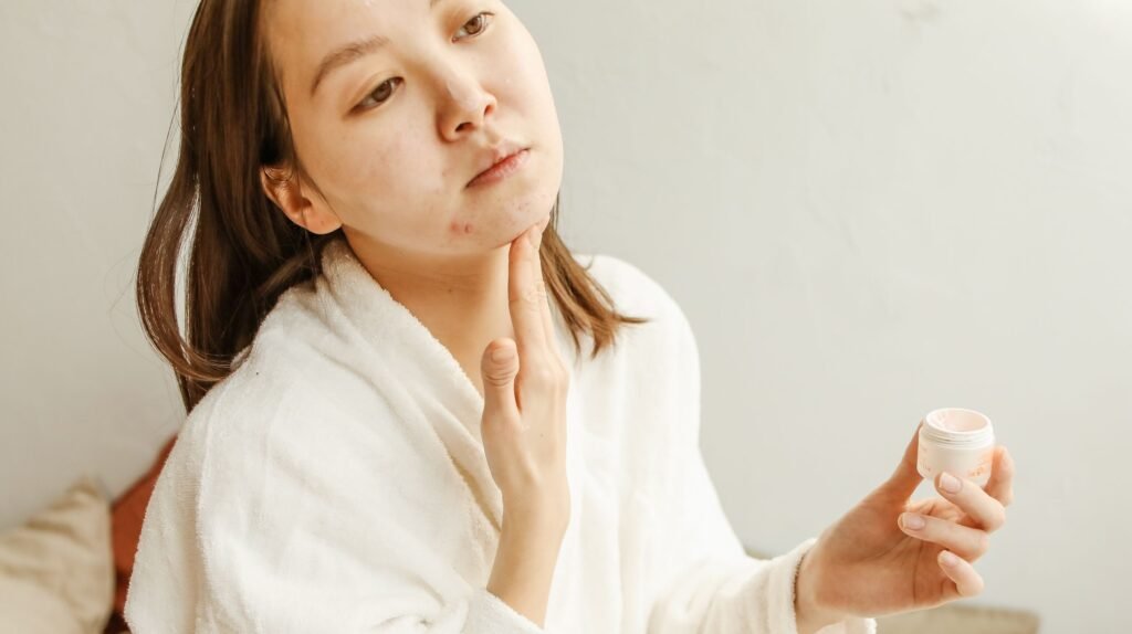 An adult woman in a bathrobe applies facial cream during her skincare routine indoors.